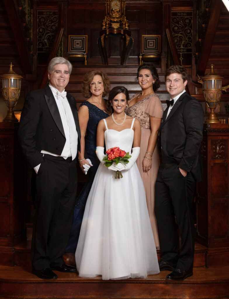 Five people in formal attire pose indoors on a staircase, with the woman in front wearing a white wedding dress and holding flowers.
